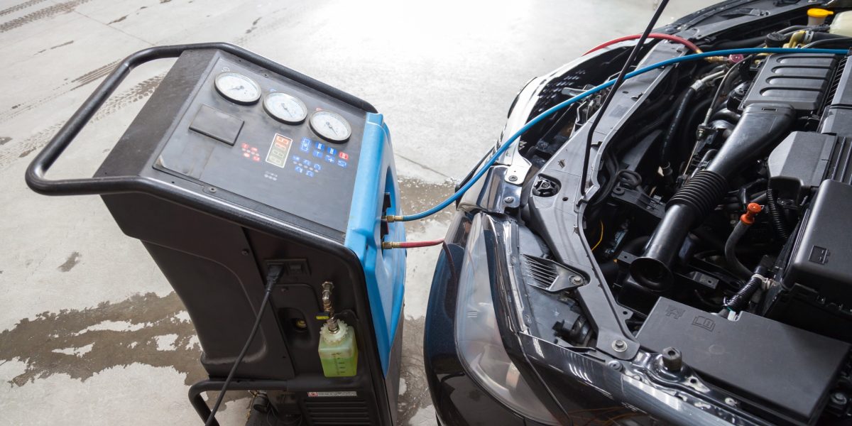 The procedure for refueling a car air conditioner using a special device with a blue compressor and a red hose near the open hood.